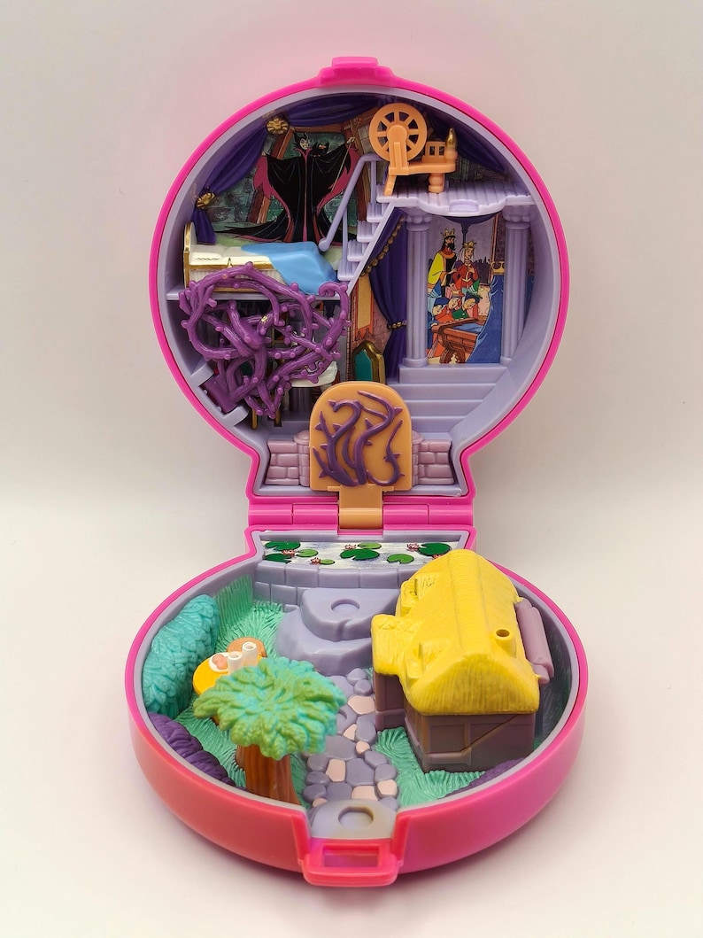 May include: A vintage pink and purple miniature playset, with a detailed interior including a bed, stairs, and a spinning wheel. The exterior features a yellow building, a small garden, and a stone path. The playset is open, revealing colourful illustrations.