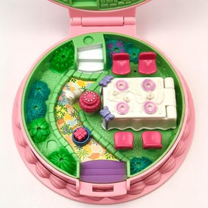May include: A vintage pink and green miniature playset, open to reveal a garden scene with a winding path, a table with pink chairs, and a small house. The playset is round with a scalloped edge.
