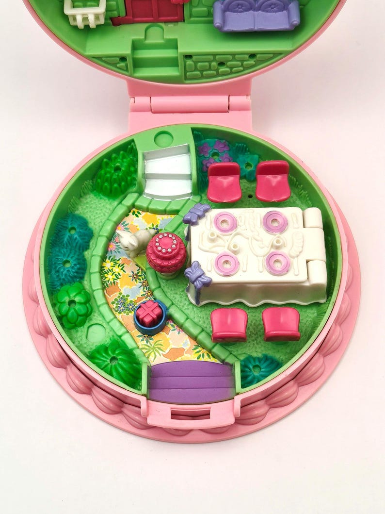 May include: A vintage pink and green miniature playset, open to reveal a garden scene with a winding path, a table with pink chairs, and a small house. The playset is round with a scalloped edge.