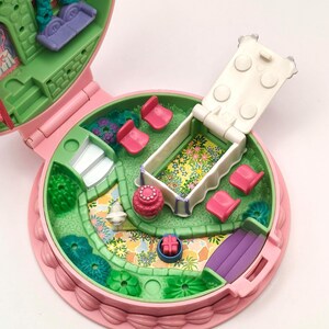 May include: A vintage pink and green miniature playset, open to reveal a garden scene. Features include a purple couch, pink chairs, a white bed with floral bedding, and a small slide. The playset is round with a scalloped edge.