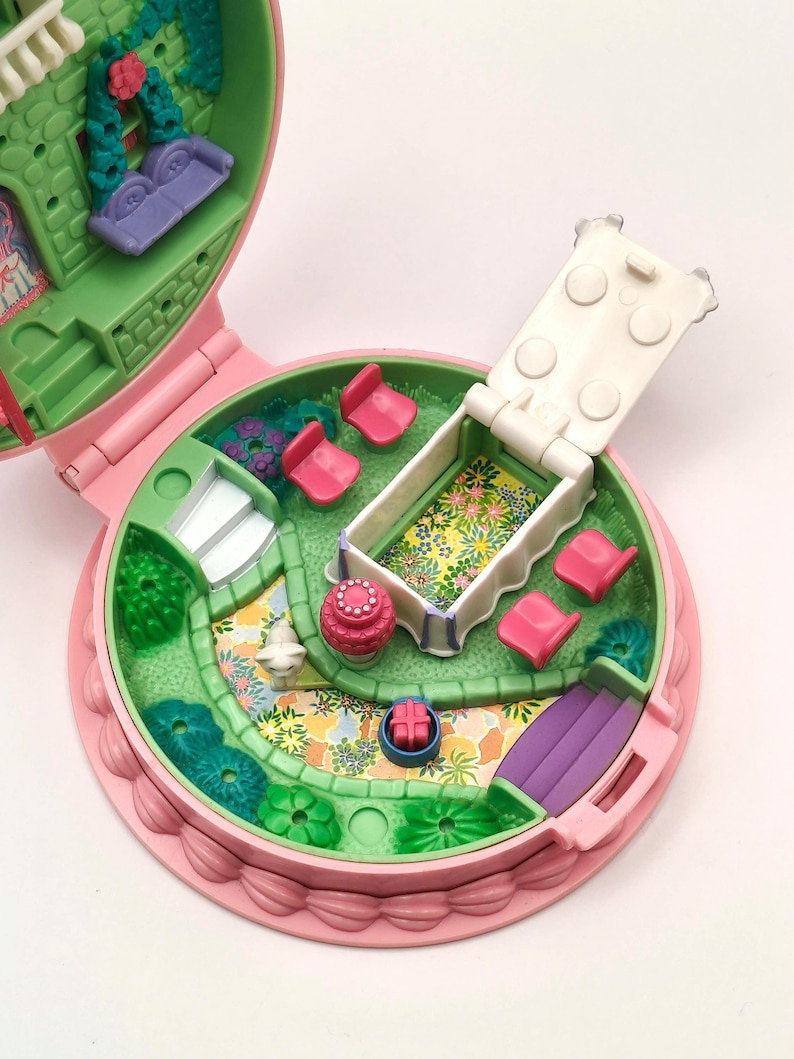 May include: A vintage pink and green miniature playset, open to reveal a garden scene. Features include a purple couch, pink chairs, a white bed with floral bedding, and a small slide. The playset is round with a scalloped edge.