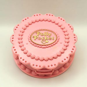 May include: A pink Polly Pocket compact in the shape of a cake. The compact has a scalloped edge and is decorated with frosting details. The gold text "Polly Pocket" is in the center, with a heart symbol.