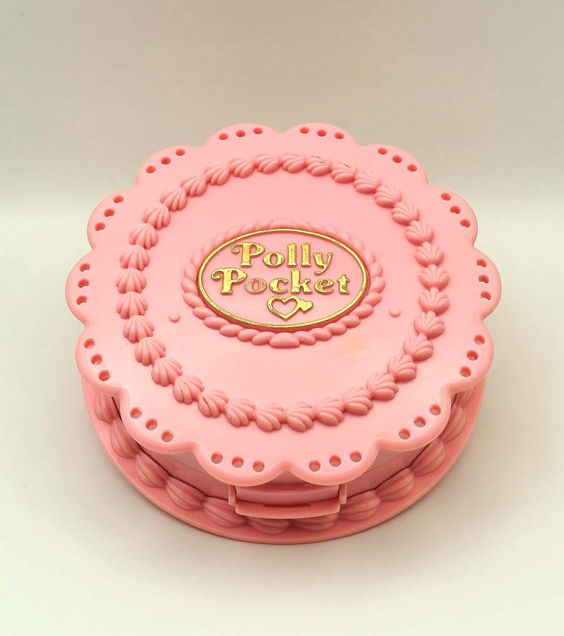 May include: A pink Polly Pocket compact in the shape of a cake. The compact has a scalloped edge and is decorated with frosting details. The gold text "Polly Pocket" is in the center, with a heart symbol.
