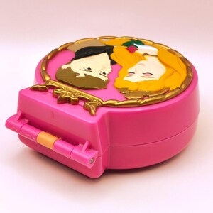 May include: A pink, heart-shaped toy compact with a gold-coloured border and a relief of two cartoon figures. The compact has a hinged opening and a small orange handle. The figures are a man with brown hair and a woman with long blonde hair.