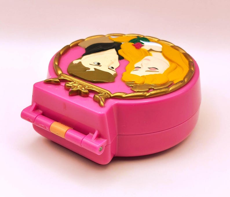 May include: A pink, heart-shaped toy compact with a gold-coloured border and a relief of two cartoon figures. The compact has a hinged opening and a small orange handle. The figures are a man with brown hair and a woman with long blonde hair.