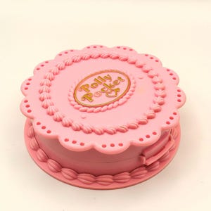 May include: A pink Polly Pocket compact toy, shaped like a cake, with a scalloped edge and decorative frosting details. The compact features the words "Polly Pocket" in gold lettering on the top.