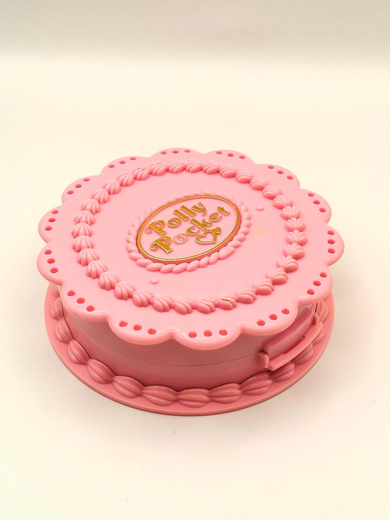 May include: A pink Polly Pocket compact toy, shaped like a cake, with a scalloped edge and decorative frosting details. The compact features the words "Polly Pocket" in gold lettering on the top.