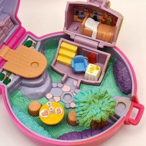 May include: A vintage, open, pink and purple miniature playset with a fairy tale theme. The playset features a bedroom, a living room, and a garden with a tree and a table. The interior includes a bed, a chair, and a table with food.