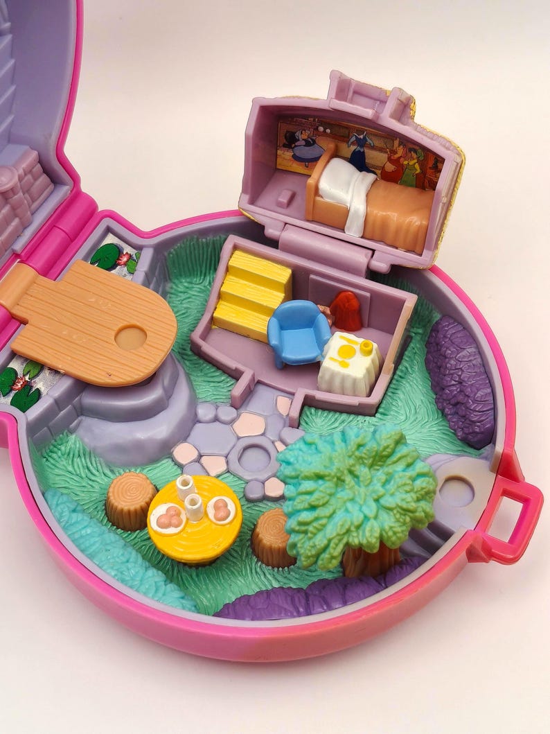May include: A vintage, open, pink and purple miniature playset with a fairy tale theme. The playset features a bedroom, a living room, and a garden with a tree and a table. The interior includes a bed, a chair, and a table with food.
