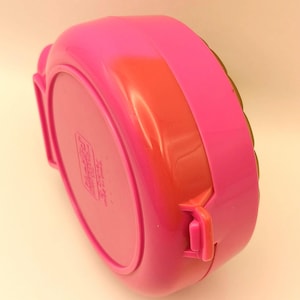 May include: A vibrant pink plastic lunchbox with a rounded rectangular form. It features an orange-red band and a pink latch. The base of the lunchbox has a printed label. The lunchbox is designed for carrying food.