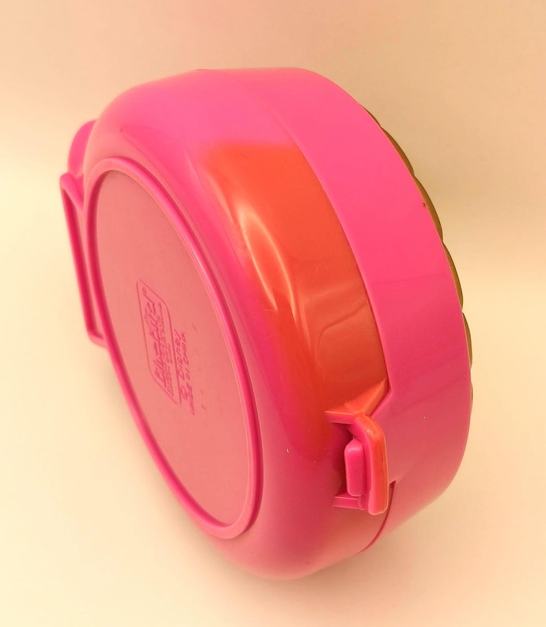 May include: A vibrant pink plastic lunchbox with a rounded rectangular form. It features an orange-red band and a pink latch. The base of the lunchbox has a printed label. The lunchbox is designed for carrying food.