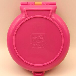 May include: A vintage, hot pink Bluebird Disney compact toy. The round, hinged case has a circular design with a raised border. The text "Bluebird" and "Disney" are visible on the bottom of the case. Made in China.