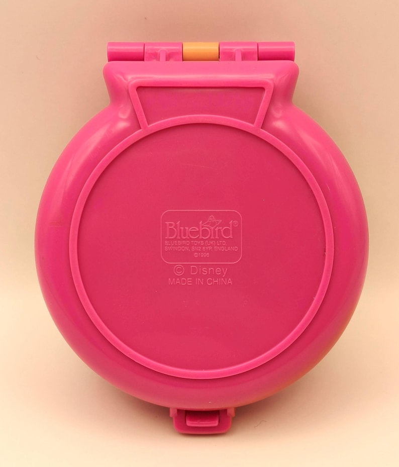 May include: A vintage, hot pink Bluebird Disney compact toy. The round, hinged case has a circular design with a raised border. The text "Bluebird" and "Disney" are visible on the bottom of the case. Made in China.