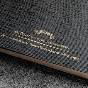K&ouml;nnte beinhalten: Schwarzes Leder-Notizbuch mit goldener Beschriftung "CALEN LEATHER CO. 400/14,8x21 cm/Blank/Made in Turkey This notebook uses Tomoe River 52g/m&sup2; white paper"