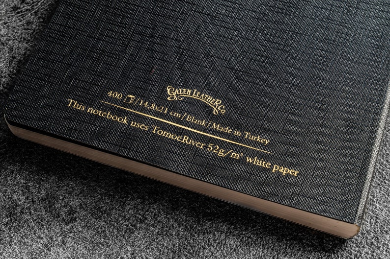 K&ouml;nnte beinhalten: Schwarzes Leder-Notizbuch mit goldener Beschriftung "CALEN LEATHER CO. 400/14,8x21 cm/Blank/Made in Turkey This notebook uses Tomoe River 52g/m&sup2; white paper"