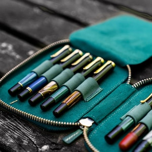 May include: A teal zippered case with multiple slots for holding pens. The case is open and filled with a variety of pens with different colored barrels and gold accents.