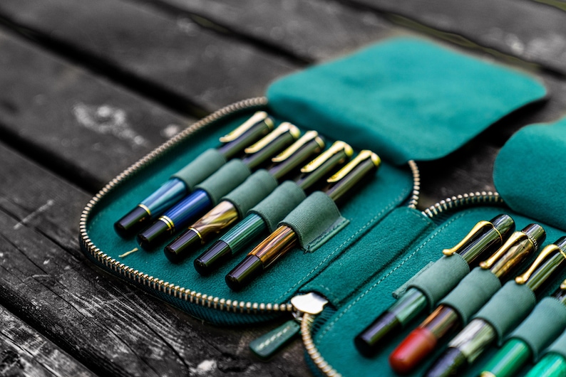 May include: A teal zippered case with multiple slots for holding pens. The case is open and filled with a variety of pens with different colored barrels and gold accents.
