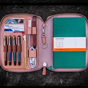 Peut inclure: Un porte-documents en cuir marron clair zippé avec un carnet Moleskine vert, quatre stylos, une règle, un crayon et un câble de recharge. Le porte-documents comporte plusieurs poches et fentes pour l'organisation.
