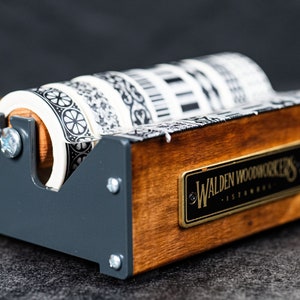 May include: A wooden tape dispenser with a black metal frame and four rolls of black and white patterned tape. The dispenser has a brass plaque that reads "Walden Woodworkers - Established".