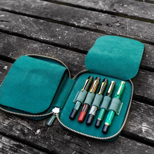 May include: A teal zippered leather case with six pens inside. The case is open and the pens are visible. The pens are different colors and have gold accents.