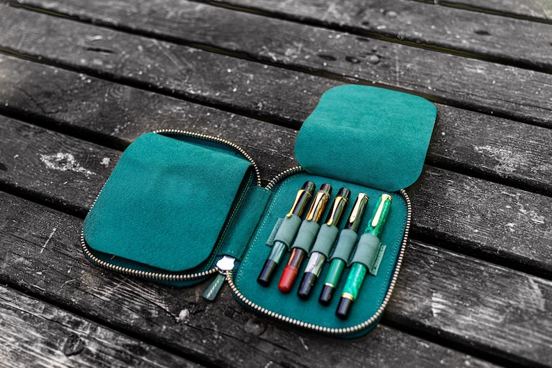May include: A teal zippered leather case with six pens inside. The case is open and the pens are visible. The pens are different colors and have gold accents.