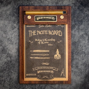 Può includere: Una lavagna in legno con cornice marrone scuro e dettagli in ottone. La lavagna presenta il testo "THE NOTE BOARD" e illustrazioni di penne e un calamaio. È presente anche il testo "Writing is the printing of the voice".