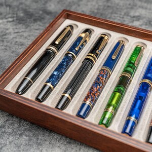 May include: A collection of fountain pens in a wooden display case. The pens feature various colors and patterns, including black, blue, green, and red. Each pen has gold-colored accents and a unique design. The case has a light gray interior.