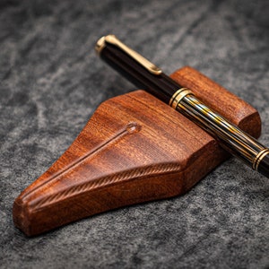 Puede incluir: Un portaplumas de madera con un acabado marr&oacute;n oscuro y una pluma estilogr&aacute;fica de rayas negras y doradas descansando encima.