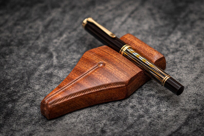Puede incluir: Un portaplumas de madera con un acabado marr&oacute;n oscuro y una pluma estilogr&aacute;fica de rayas negras y doradas descansando encima.