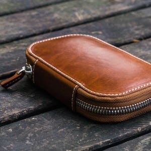 May include: A brown leather zippered pouch with white stitching. The pouch is lying on a wooden surface.