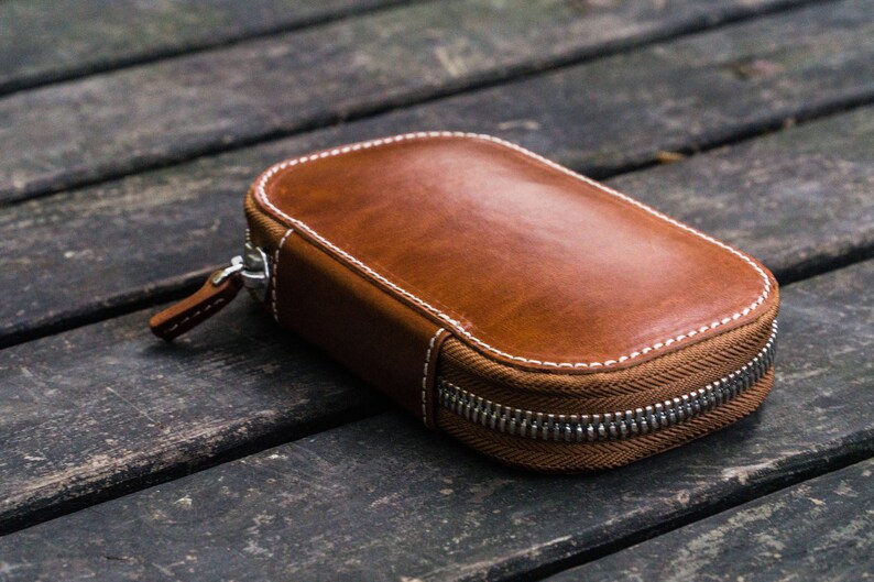 May include: A brown leather zippered pouch with white stitching. The pouch is lying on a wooden surface.
