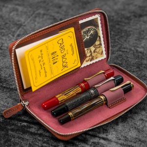May include: A brown leather zipped case with a pink interior. The case contains three pens and a yellow card with the text "The Card Book" and "Billa" written on it.