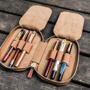 May include: A brown leather pen case with a zipper closure, open to reveal six pens of various colors and styles.