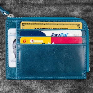 May include: A teal leather wallet with a zipper closure, holding several cards. Visible cards include an American Express, a PayPal card, and a G bonus card. The wallet is open, displaying the card slots.