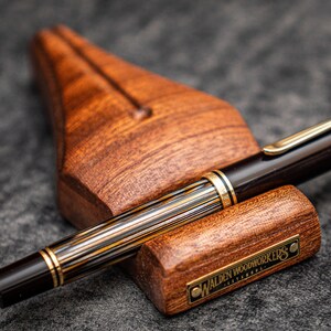 Puede incluir: Un portaplumas de madera marr&oacute;n con una placa de metal dorado que dice "Walden Woodworkers Estambul" y una pluma estilogr&aacute;fica de rayas negras y doradas descansando encima.