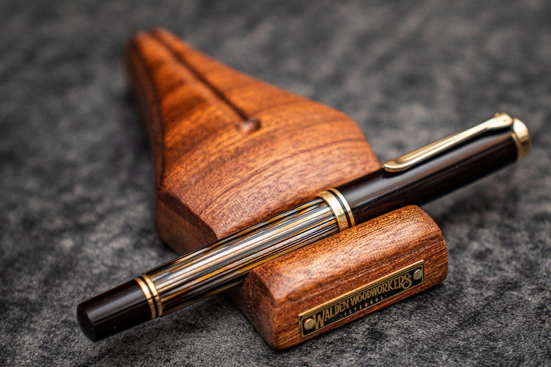 Puede incluir: Un portaplumas de madera marr&oacute;n con una placa de metal dorado que dice "Walden Woodworkers Estambul" y una pluma estilogr&aacute;fica de rayas negras y doradas descansando encima.