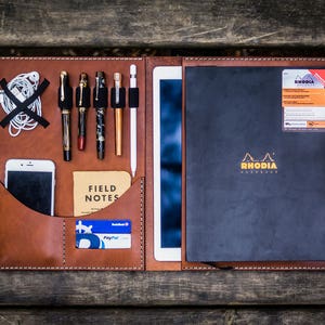 May include: A brown leather portfolio with a black notebook, a white tablet, a smartphone, pens, and a Field Notes notebook. The notebook has the Rhodia logo and the text "RHODIA" on the cover.
