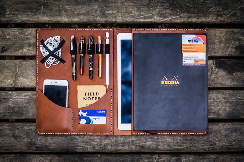 May include: A brown leather portfolio with a black notebook, a white tablet, a smartphone, pens, and a Field Notes notebook. The notebook has the Rhodia logo and the text "RHODIA" on the cover.