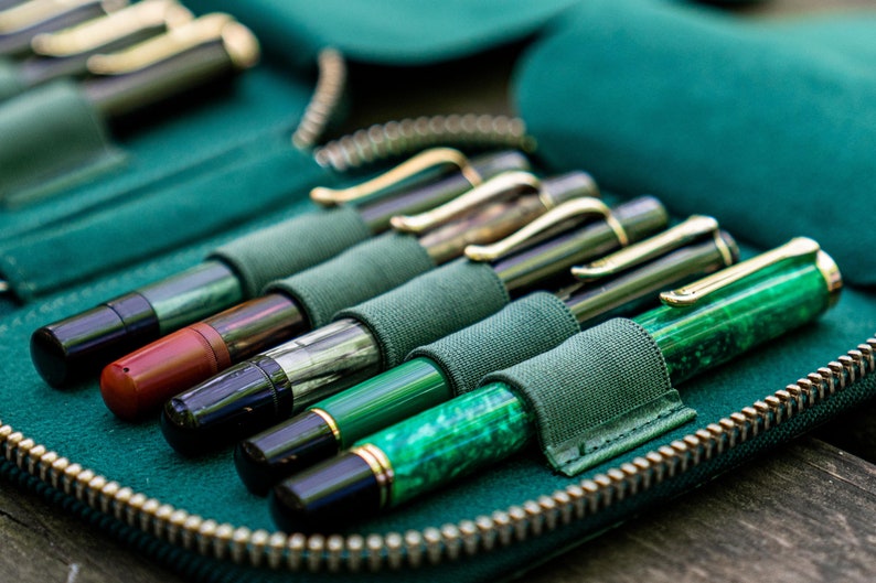 May include: A green leather pen case with a zipper closure, holding a collection of fountain pens with different colored barrels and nibs.