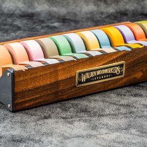 May include: A wooden tape dispenser with a black metal base and a label that reads "Walden Woodworkers - Istanbul". The dispenser holds 10 rolls of colorful tape.