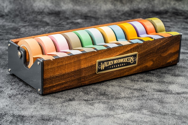 May include: A wooden tape dispenser with a black metal base and a label that reads "Walden Woodworkers - Istanbul". The dispenser holds 10 rolls of colorful tape.