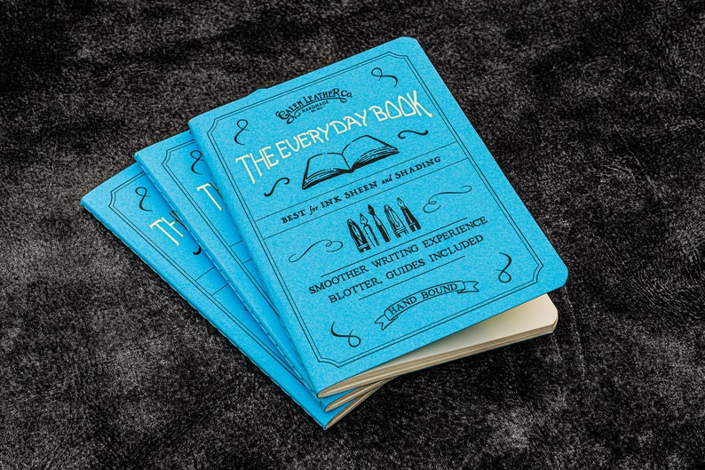 Pu&ograve; includere: Tre quaderni blu impilati uno sopra l'altro. Il quaderno in cima ha il testo "The Everyday Book" in lettere nere. Il testo "Best for Ink Sheen and Shading" &egrave; anche stampato sulla copertina anteriore. Il quaderno &egrave; rilegato a mano.