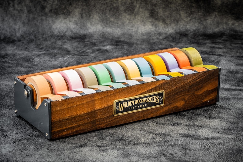 May include: A wooden tape dispenser with a black metal base and a label that reads "Walden Woodworkers - Istanbul". The dispenser holds ten rolls of colorful tape.