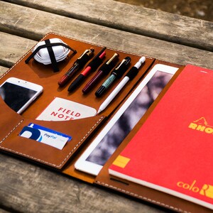 May include: A brown leather portfolio with a zipped closure, containing a tablet, a smartphone, a notebook, pens, and other accessories. The notebook is red and has the brand name "RHODIA" printed on the cover.