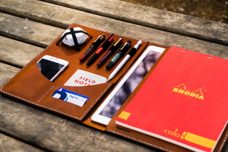 May include: A brown leather portfolio with a zipped closure, containing a tablet, a smartphone, a notebook, pens, and other accessories. The notebook is red and has the brand name "RHODIA" printed on the cover.