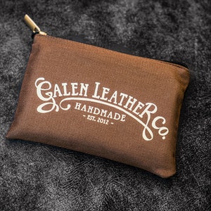 Pu&ograve; includere: Una pochette in tela marrone con una chiusura a zip. La pochette ha un logo bianco che recita "Galen Leather Co. Handmade - Est. 2012 - "