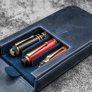 Op de afbeelding: Een marineblauwe leren pennenetui met drie pennen. De pennen zijn zwart, rood en oranje met gouden details. De etui is open en toont een donkerblauwe binnenkant. De etui ligt op een getextureerd grijs oppervlak.