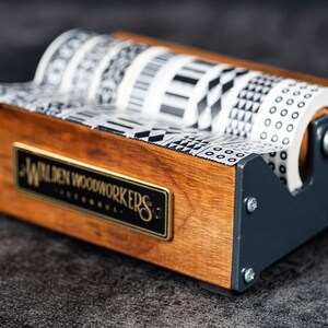 May include: Wooden tape dispenser with a black and white patterned tape roll. The dispenser has a metal base and a wooden top with the text "Walden Woodworkers" engraved on it.