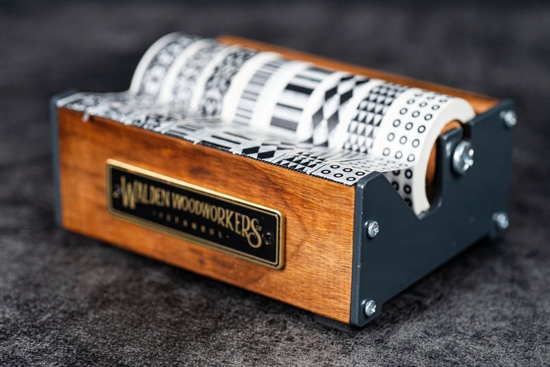 May include: Wooden tape dispenser with a black and white patterned tape roll. The dispenser has a metal base and a wooden top with the text "Walden Woodworkers" engraved on it.