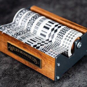 May include: A wooden and metal tape dispenser with six rolls of black and white patterned washi tape. The dispenser has a label that reads "Walden Woodworkers".